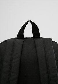 Sac à dos noir avec bretelles rembourrées et une boucle de portage en tissu tissé noir, sur un fond clair uni.