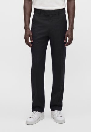 Homme portant un pantalon formel noir et des baskets blanches, debout devant un fond clair uni.
