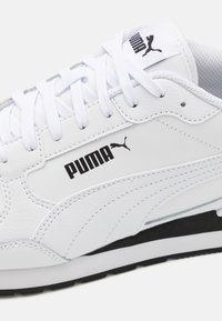 Baskets blanches en cuir avec des accents noirs. Elles présentent un col rembourré, un logo bien visible et une semelle texturée pour une meilleure adhérence. Les lacets sont blancs.