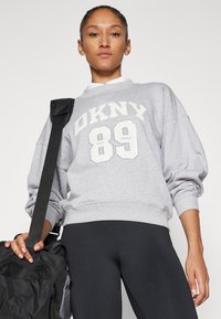 Pilkas sportinis džemperis su balta "DKNY 89" užrašu, su rausvomis rankovėmis ir apačia. Suderintas su juodomis tampomis ir juoda kuprine su dirželiu.