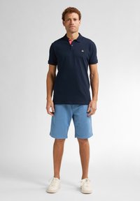 Marinblå pikétröja med röda krageaccent och logotyp, tillsammans med ljusblå shorts. Har korta ärmar och en klassisk passform. Vita sneakers fulländar looken.