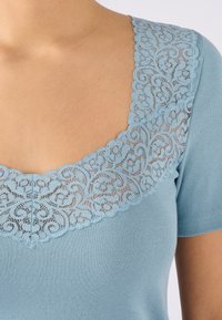 Maglietta azzurra chiara a maniche corte con scollatura in pizzo a onde e motivi floreali, texture liscia e design aderente.