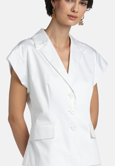 Femeie care poartă un sacou alb fără mâneci, cu rever decupat și trei nasturi în față, accesorizată cu cercei aurii filigranați.