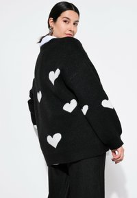 Schwarzer übergroßer Pullover mit weißen Herzmustern, gestrickter Textur und fallenden Schultern. Über einem Kragenhemd getragen, mit geripptem Saumdetail.