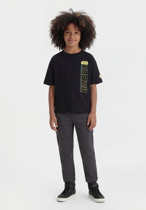 Garçon aux cheveux bouclés portant un t-shirt noir Star Wars Vader, un pantalon cargo gris et des baskets montantes noires, debout sur fond blanc.