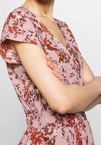 Kukkamekko pehmeästä vaaleanpunaisesta kankaasta, jossa on punaisia ja ruskeita kukkakuoseja, V-mallinen kaula-aukko ja lyhyet hihat. Varustettu joustavalla vyötäröllä, joka antaa istuvan ilmeen.