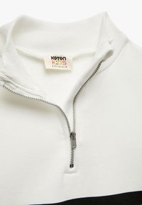 Valkoinen vetoketjullinen huppari, jossa on ribatty kaulus ja musta vaakasuora raita alaosassa. Mukana hopeinen vetoketju ja tarra, jossa lukee "Koton Kids."