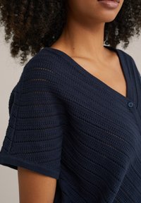 Jūras zilās krāsas adīta kardigāna/vēlības jaku ar V veida kaklu, ar svītrainu tekstūru un īsām piedurknēm. Priekšā ar vienu pogu, lai aizvērtu.