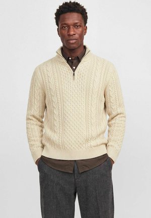 Un homme porte un pull crème en maille torsadée à demi-zip sur une chemise marron et un pantalon foncé, sur un fond blanc uni.