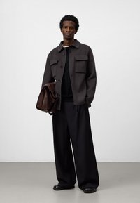 Ciemnoszary wełniany żakiet z dwiema kieszeniami na piersi, czarny sweter pod spodem, luźne czarne spodnie i brązowa skórzana torba na ramię.