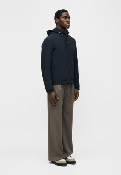 Άνδρας όρθιος, φοράει μπλε ναυτικό μπουφάν με κουκούλα, φαρδιά καφέ παντελόνια και μπεζ παπούτσια με μαύρες σόλες, σε ένα μονόχρωμο φωτεινό φόντο.