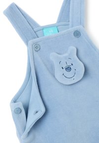 Overoles de bebé en un suave color azul claro con tirantes abotonados y un parche de cara de oso sonriente en el pecho, confeccionados en tela suave.