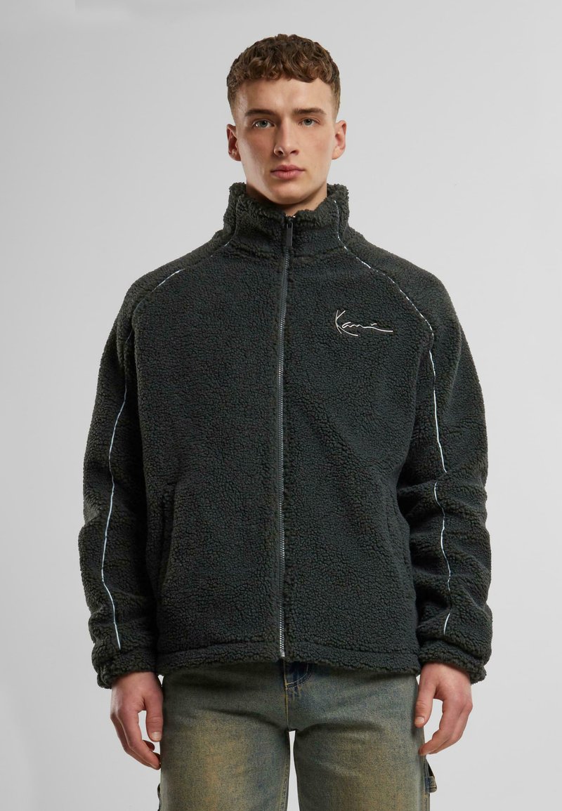 Fleece zip-up jacket in dark green with a soft texture, contrasting white stitching, stand-up collar, and branded embroidery on the chest.