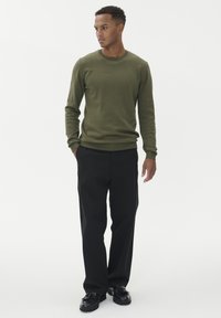 Olivově zelený svetr s dlouhým rukávem a žebrovanými manžetami, v kombinaci s černými kalhotami rovného střihu a černými botami, na jednobarevném bílém pozadí.