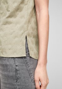 Blouse vert clair avec motifs en relief en forme d'éventail, fentes latérales, et finition texturée. Associée à un jean gris pour un look superposé.