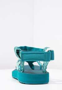 Sandal con cinturino regolabile color teal, con suola texturizzata, vista da dietro su una superficie e sfondo bianchi.