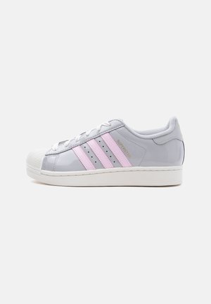 Biela a sivá teniska Adidas Superstar s ružovými pruhmi, bielymi šnúrkami a hrubou bielou podrážkou, zobrazená z bočného pohľadu na jednoduchom pozadí.