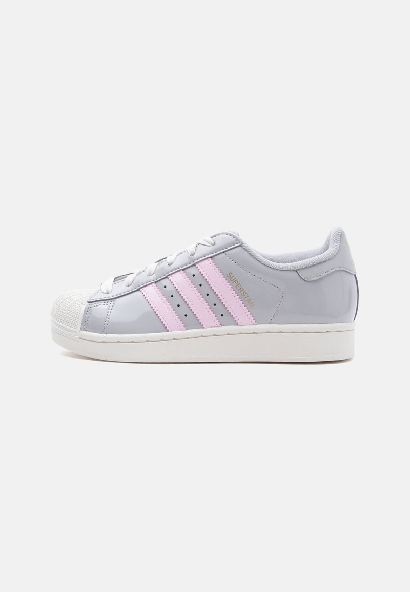 Λευκό και γκρι παπούτσι Adidas Superstar με ροζ ρίγες, λευκά κορδόνια και παχύ λευκό πάτο, εμφανιζόμενο από πλάγια όψη σε απλό φόντο.
