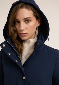 Chaqueta azul marino con capucha, cierre delantero con cremallera y detalles de herrajes dorados. Se lleva sobre un suéter beige claro con un patrón texturizado.