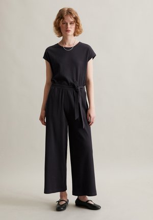 Femme aux cheveux courts et bouclés portant une combinaison noire côtelée avec une ceinture nouée à la taille et des chaussures plates noires, debout devant un fond uni.