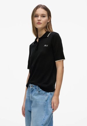 Ung kvinde iført en sort kortærmet polo med hvide kanter og lyseblå højtaljede jeans, stående foran en hvid baggrund.