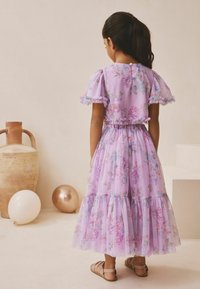 Vestido floral lila con mangas con volantes, falda de tul en capas, cintura elástica y cierre de botones en la espalda. Combinado con sandalias marrones.