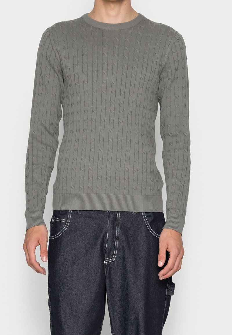 Grå kabelstickad tröja med rund halsringning och muddar i ribbstickning, tillsammans med mörka denimbyxor. Texturen visar ett upphöjt mönster och detaljerad sömnad.