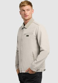 Homme portant une veste zippée beige clair avec col et pantalon noir, mains dans les poches, regardant vers la gauche sur un fond uni.