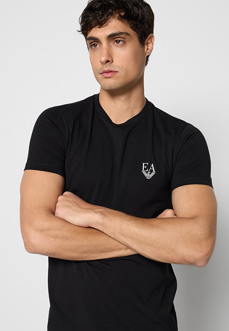 Joven con pelo corto y oscuro, con una camiseta negra con el logo de EA, de pie con los brazos cruzados frente a un fondo liso gris claro.