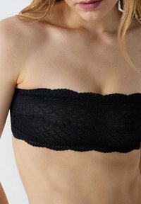 Top bandeau in pizzo nero con un motivo testurizzato e bordi ricamati, progettato per offrire una vestibilità senza spalline. Vista ravvicinata del materiale.