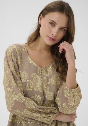 Tânără cu păr castaniu ondulat purtând o bluză bej cu model floral auriu, sprijinindu-și capul în mână, fundal neutru.