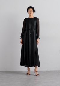 Robe noire à manches longues avec un corsage ajusté et une jupe fluide, dotée d'une taille à nouer. Fabriquée en tissu léger avec des manches transparentes.