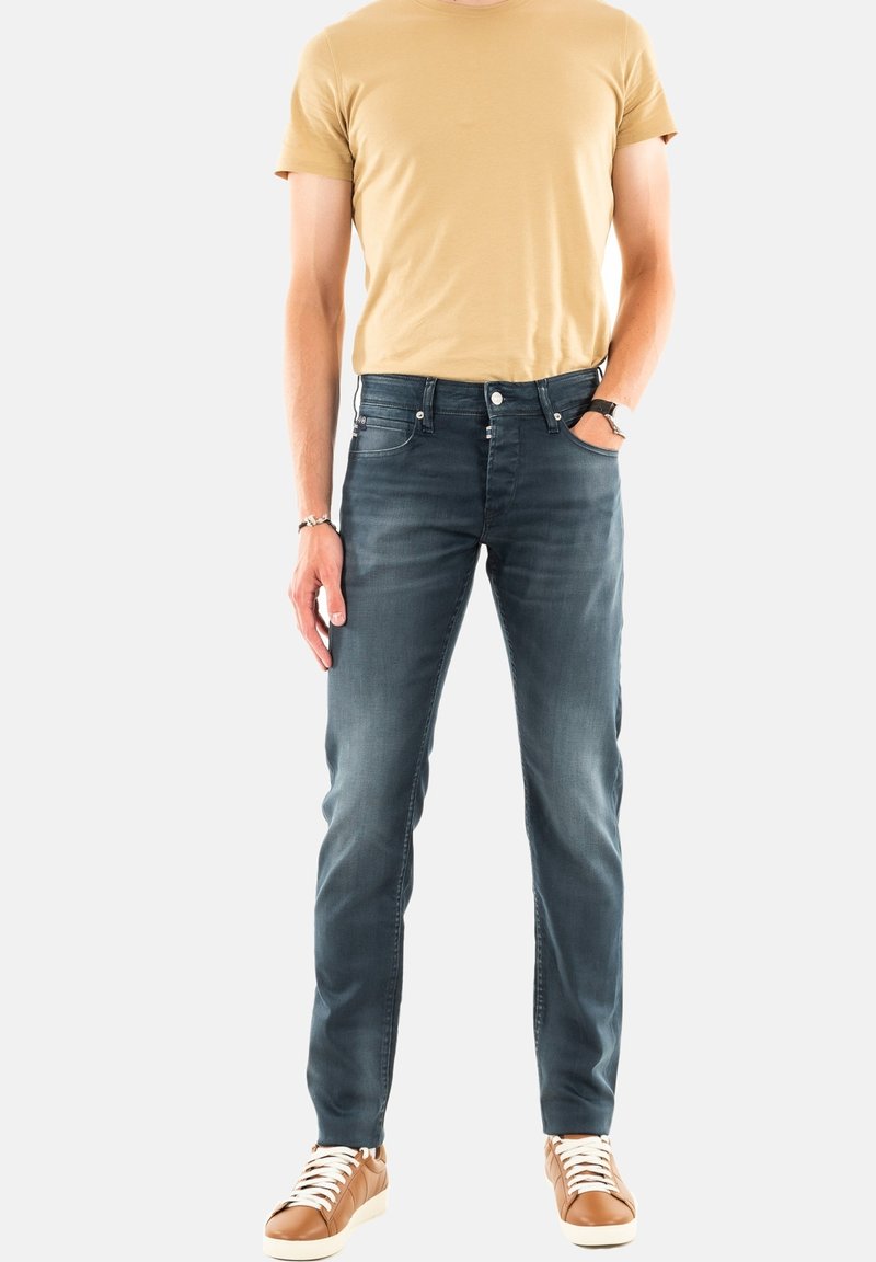 Jean slim en denim bleu foncé, avec cinq poches, une fermeture à bouton classique et un léger effet délavé. Porté avec un t-shirt beige.