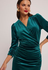 Szmaragdowa welurowa sukienka kopertowa z długimi, marszczonymi rękawami i marszczeniem w talii. Gładka tekstura, głęboki dekolt w kształcie litery V. Złote kolczyki w kształcie kół.