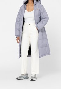 Ljuslila lång vadderad jacka med dragkedja, huva och elastiska muddar; bärs över en vit linne och cremefärgade byxor, kombinerat med grå sneakers.