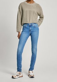 Persona con un suéter beige de punto, jeans ajustados azul de talle alto y zapatillas blancas con detalles beige, de pie frente a un fondo liso.