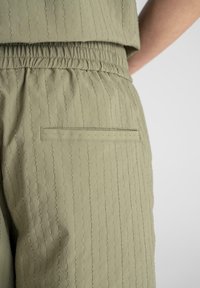 Lys olivengrønne bukser med en teksturert stoff med vertikale striper, elastisk liv, og en baklomme.
