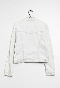 Bílá džínsová bunda visící na černém ramínku na pozadí čistě bílé stěny, viděná zezadu.