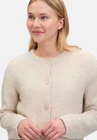 Beige ulltröja med rund halsringning, mjuk текстur, detaljer med gyllene knappar och långa ärmar. Knäppt fram med en avslappnad passform.