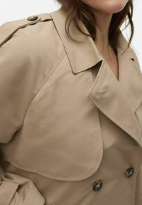 Trench-coat beige avec un large col, boutons double boutonnage et épaulettes ; tissu lisse avec un design structuré.
