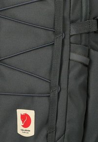 Primo piano del tessuto del zaino Fjällräven nero con trama fitta, corde elastiche e patch con il logo raffigurante la testa di una volpe rossa.