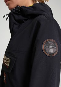 Giacca parka navy con tasca frontale, patch con logo sulla manica, cappuccio regolabile e tessuto impermeabile liscio.