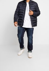 Czarna pikowana kurtka z wysokim kołnierzem, w zestawieniu z białym t-shirtem i ciemnymi dżinsami. Model nosi białe tenisówki, prezentując swobodny styl.