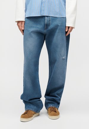 Moški, ki nosi ohlapne modre kavbojke z majhno trganino na desnem stegnenici, rjave semiš superge in modro črtasto srajco z belimi dolgimi rokavi.