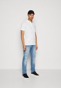 Hvid polo shirt med krave, kombineret med lyseblå slidte jeans og sorte sneakers. Modellen bærer en sort kasket.