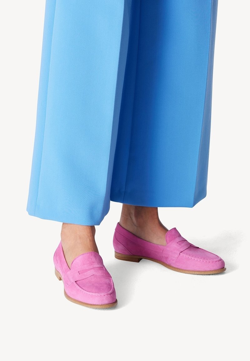 Mocassins en daim rose avec un talon plat et une sangle décorative à l'avant, associés à un pantalon large bleu. Texture lisse et design classique.