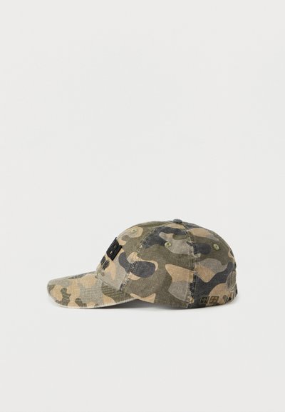 Καπέλο παραλλαγής σε πράσινο και μπεζ ύφασμα με καμπύλη γείσο, υφή επιφάνειας και κεντημένο μαύρο λογότυπο στο μπροστινό μέρος.