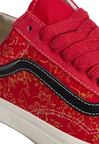 Czerwony tenisówka z płótna z złotymi kwiatowymi wzorami, czarnym paskiem po bokach i dopasowanymi czerwonymi sznurówkami. Posiada białą gumową podeszwę.