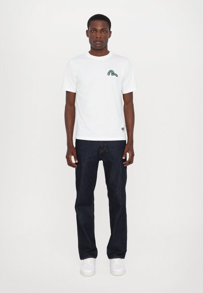 Camiseta blanca de manga corta con diseño bordado en verde en el lado izquierdo del pecho, combinada con jeans oscuros de mezclilla y zapatillas blancas.