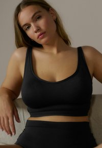 Schwarzes nahtloses Crop-Top mit breiten Trägern, weicher Textur und rundem Ausschnitt, kombiniert mit schwarzen hochtaillierten Unterteilen, präsentiert auf einem neutralen Hintergrund.